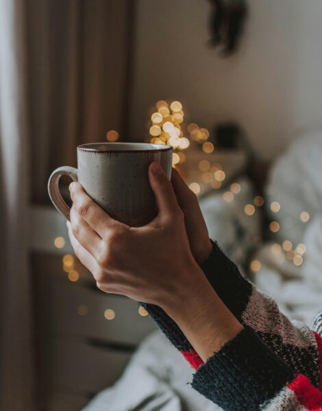 Hands holding a warm mug indoors with soft bokeh lights, creating a cozy winter atmosphere.